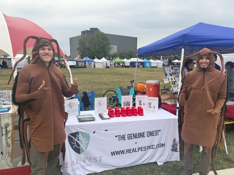 Two people in cockroach costumes stand behind a Real Pest Solutions table at an outdoor event, smiling and posing.
