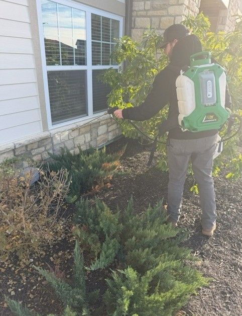 A pest control technician in dark clothing wears a green backpack sprayer while treating the exterior of a house.