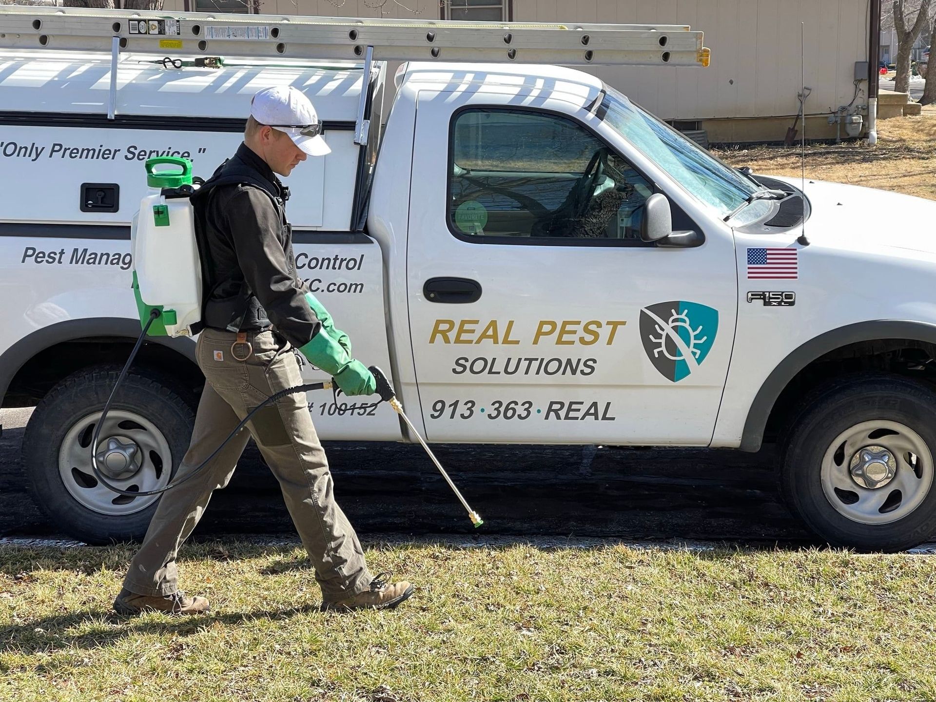 A pest control technician in a work uniform walks across a lawn carrying a sprayer beside a company truck.