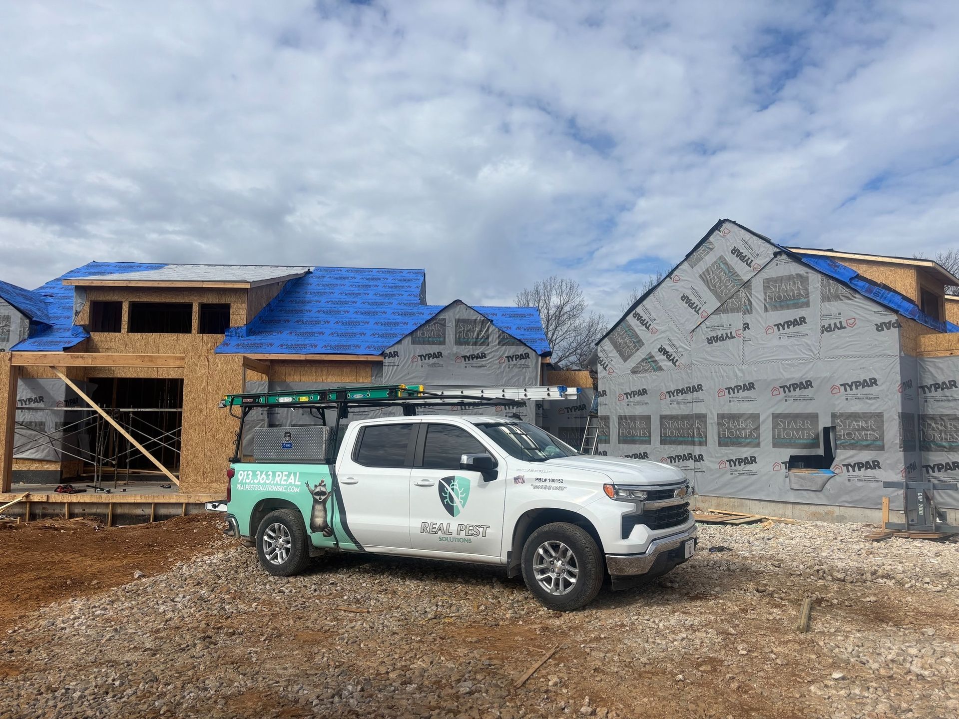 A white pickup truck with a logo parked at a residential construction site featuring wood frames and blue roofing paper.