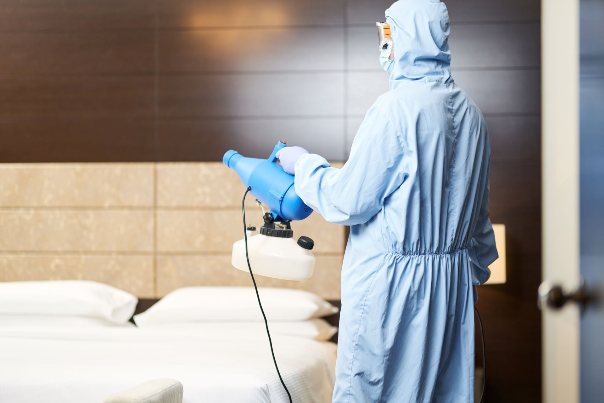 A person in a light blue protective suit uses a handheld fogger to disinfect a hotel room bed.
