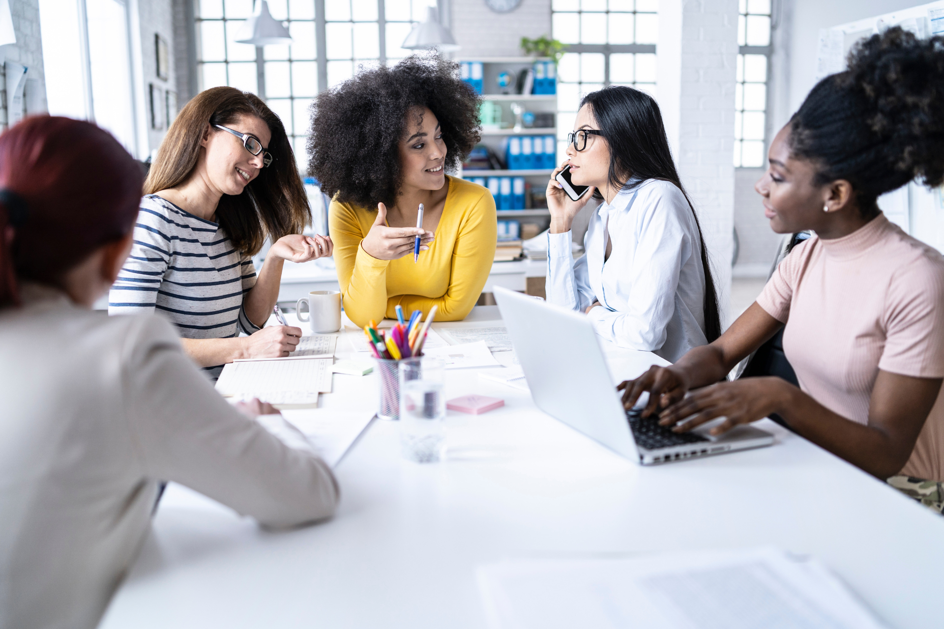 Five women at a white table in an office, discussing work with a laptop and documents.