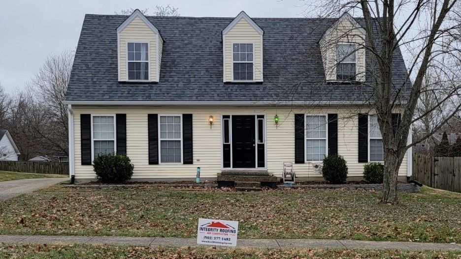 A beige two-story house with a dark gray roof, black shutters, and three dormers. A sign is in the front yard.