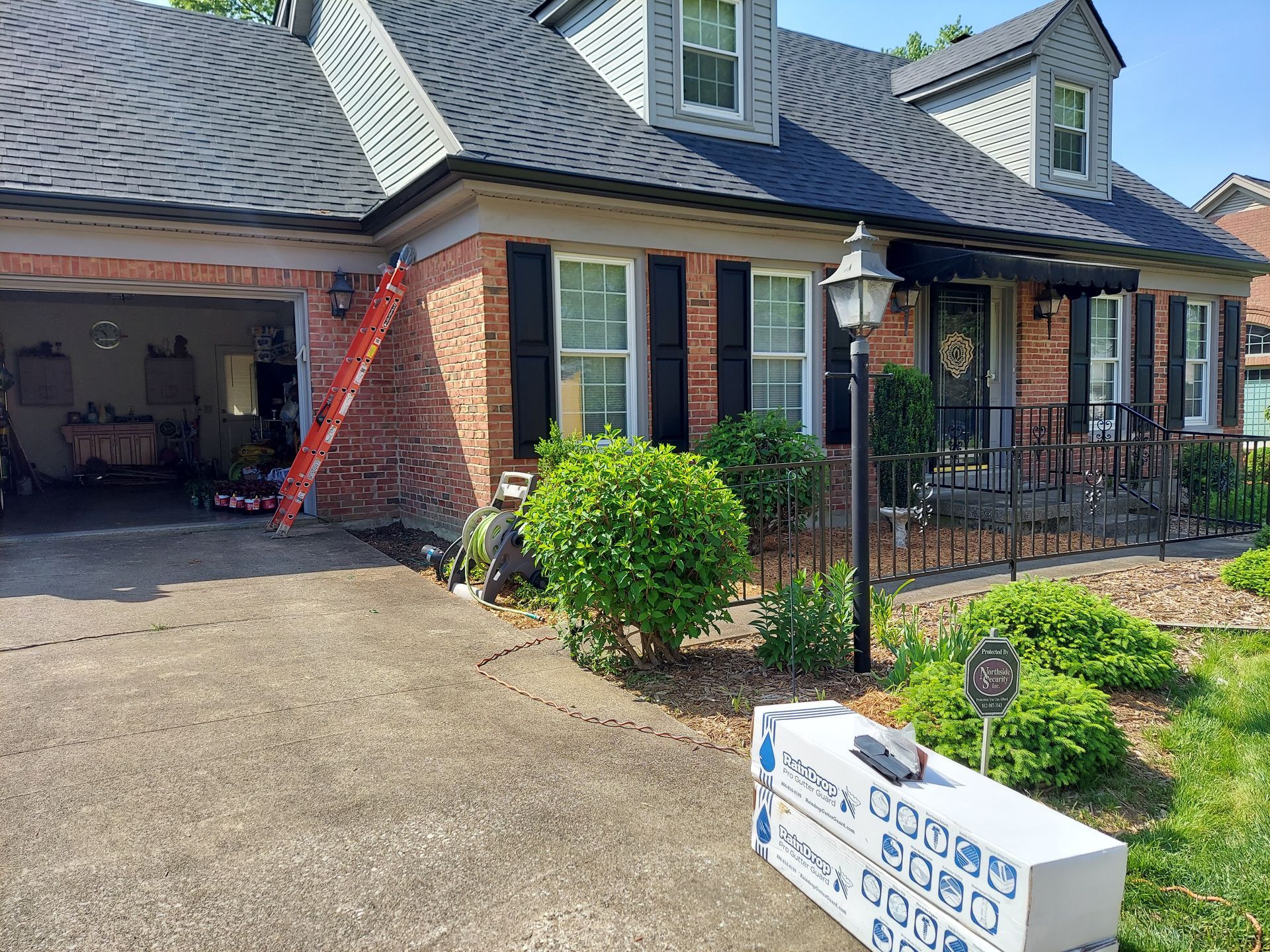 A brick house with black shutters, a dark roof, and a ladder leaning against it. A stack of materials sits on the driveway.