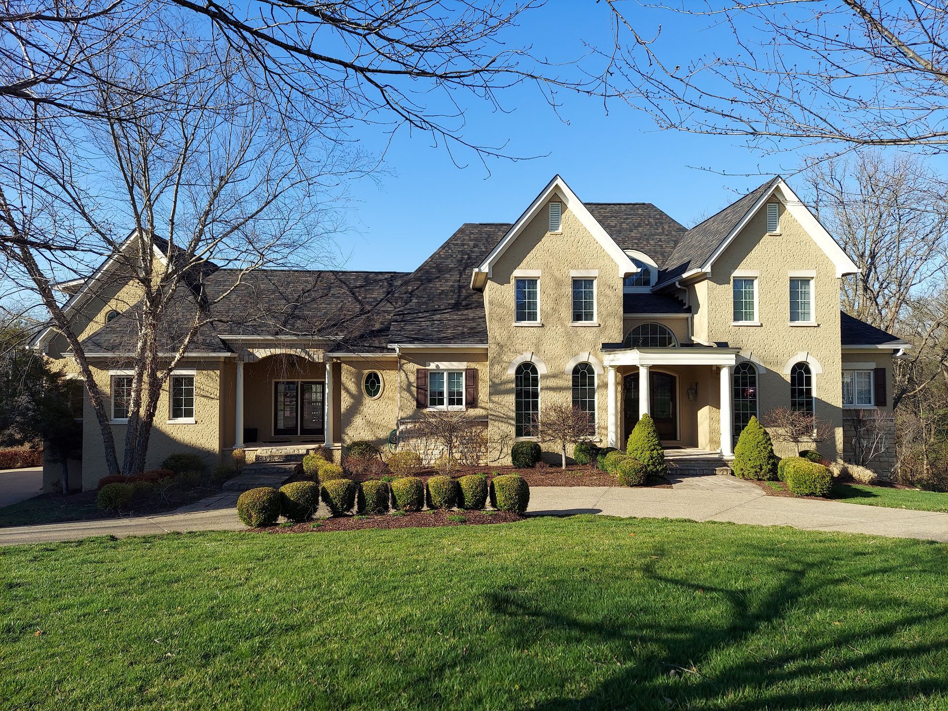 Two-story beige brick house with black roof, manicured lawn, and bare trees under a blue sky.