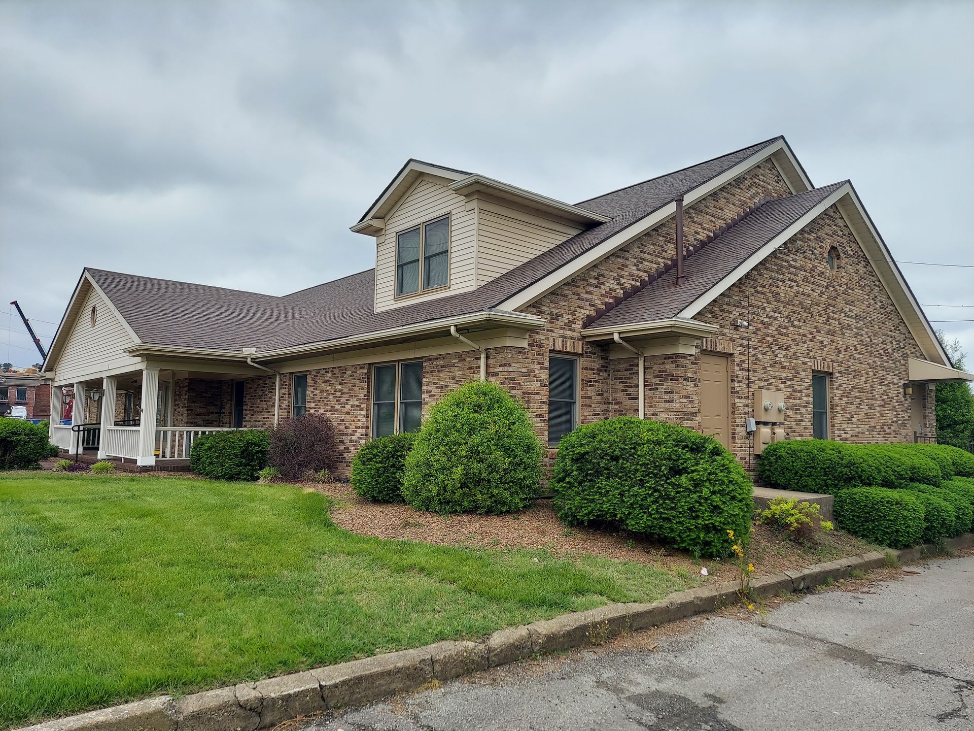 Brick building with brown roof and beige dormer, green lawn and shrubs, overcast sky.
