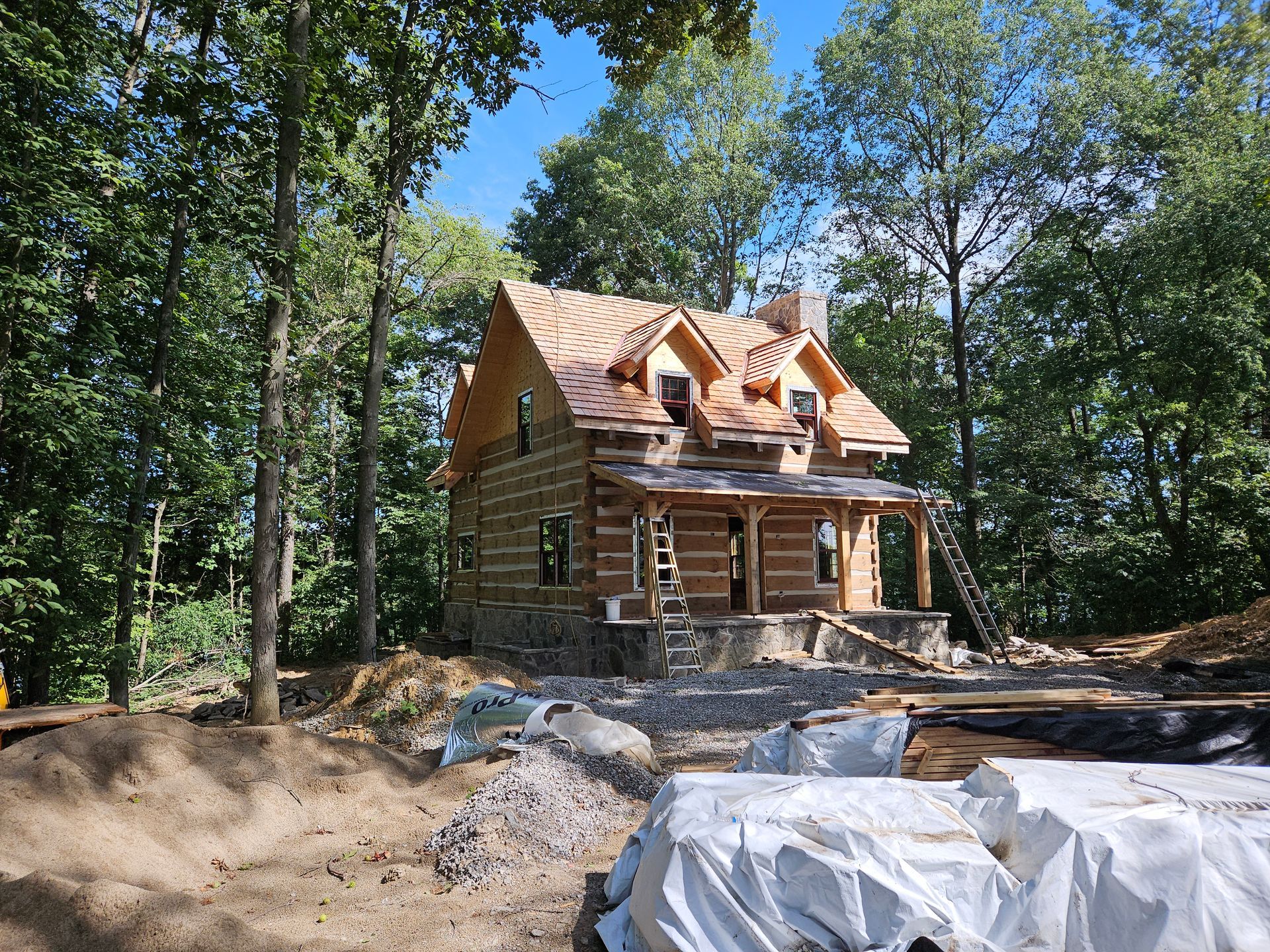 Log cabin under construction in a wooded area with exposed roof beams and stone foundation.