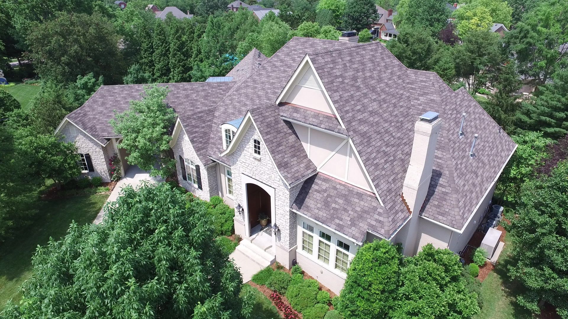 An aerial view of a large, beige house with a brown shingled roof, surrounded by green trees.