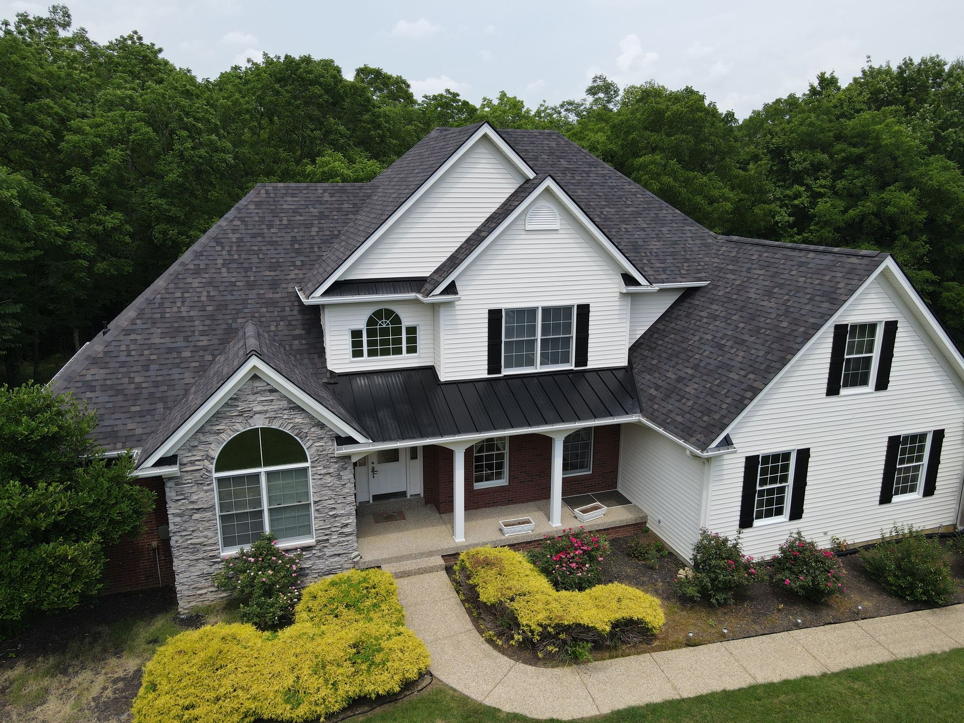 Two-story white house with dark roof and black shutters, brick and stone accents, surrounded by greenery.