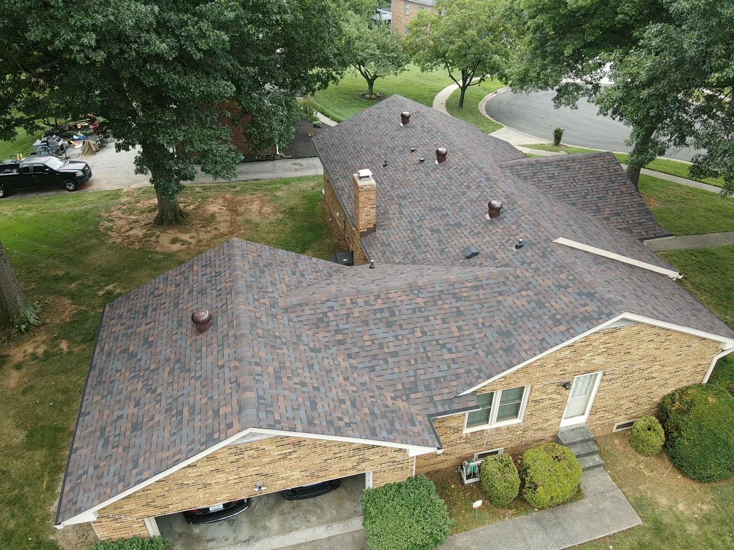 Overhead view of a house with a brown shingled roof, trees, lawn, and driveway.