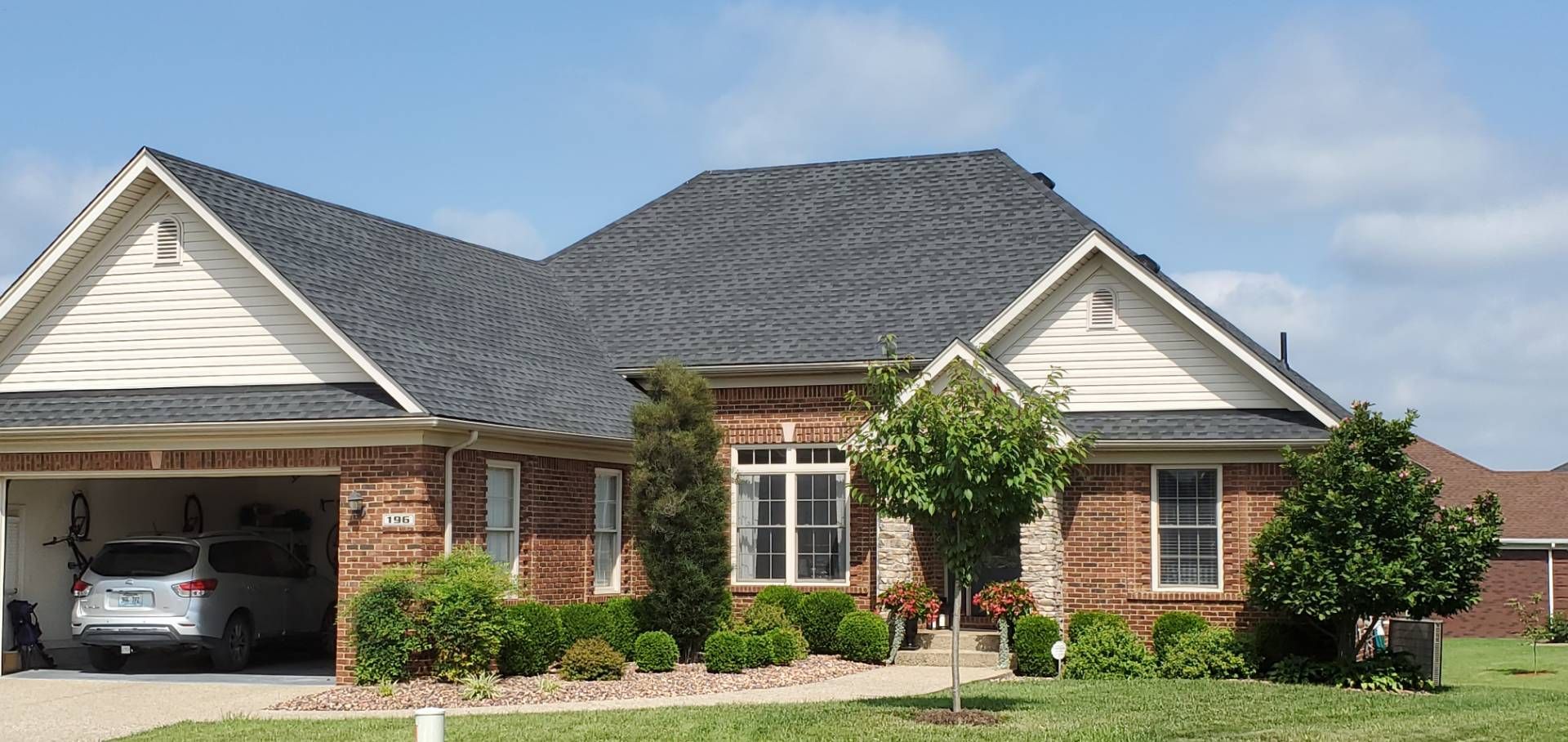 A brick house with a gray roof and a garage. Green bushes and a lawn in front. Blue sky.