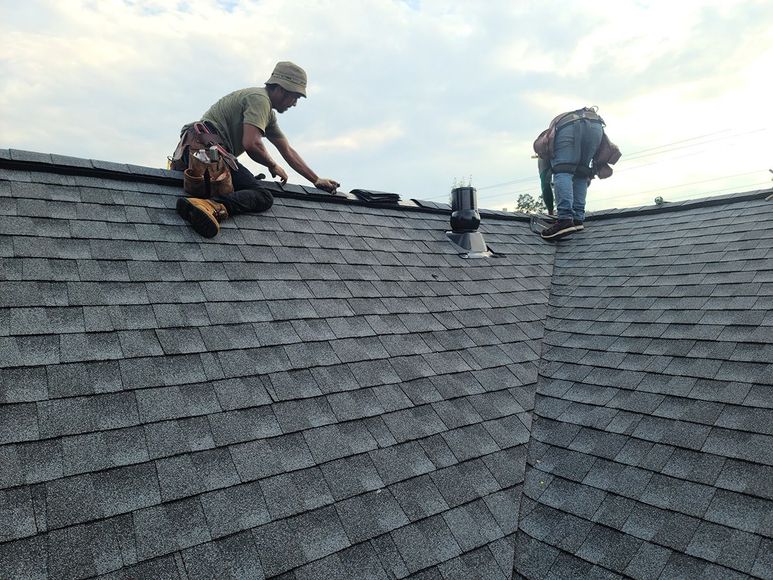Two roofers installing shingles on a gray asphalt shingled roof, cloudy sky.