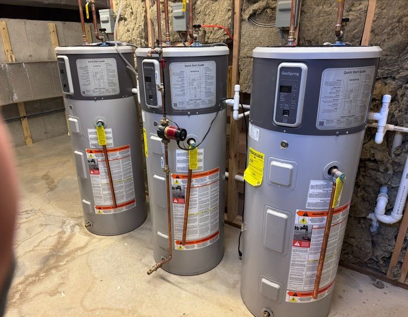 Three gray water heaters in a basement, pipes and wiring visible.