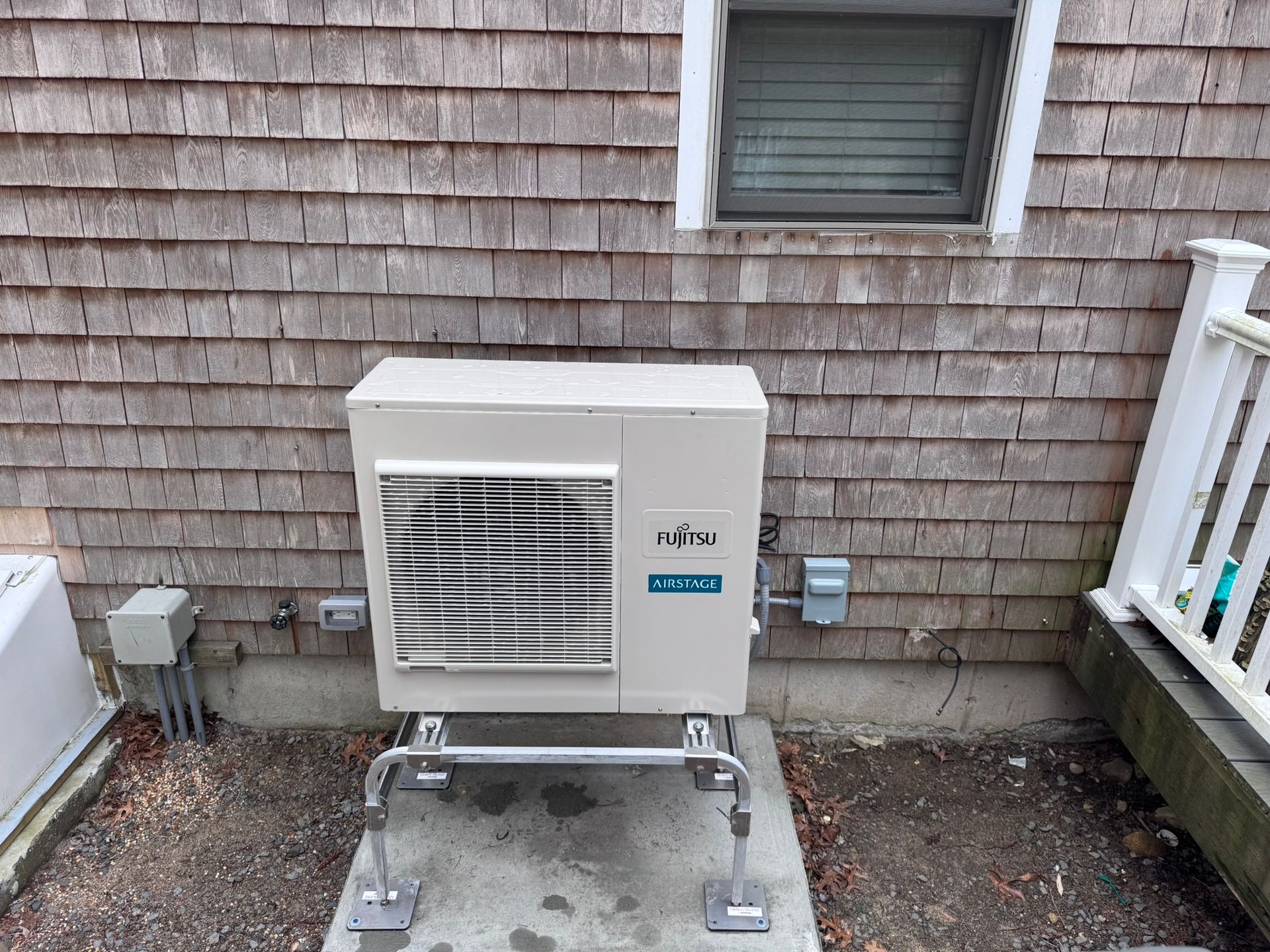 Fujitsu heat pump unit mounted on a concrete slab, installed outside a wood-shingled house.