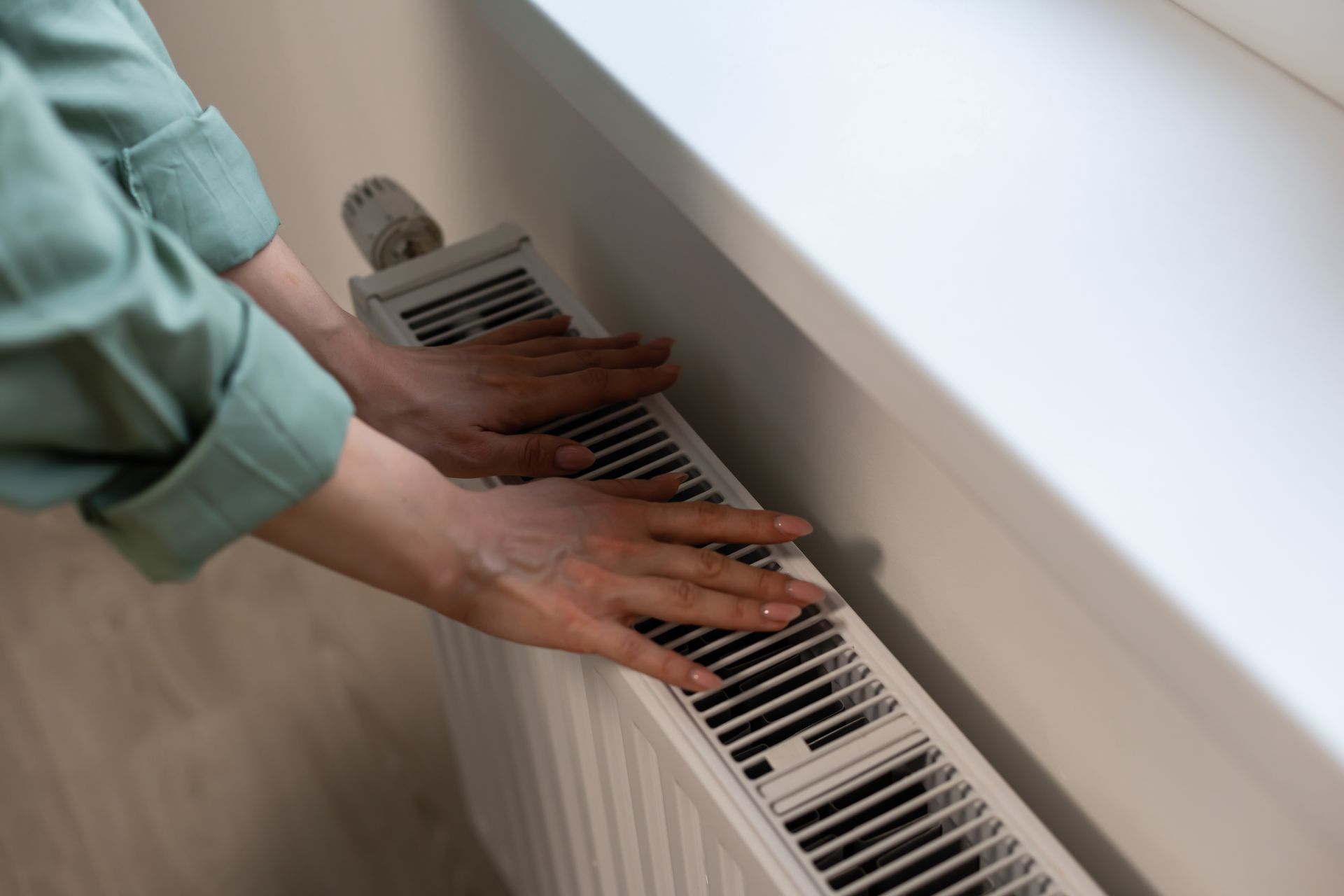 Hands touching a white radiator near a window, checking for heat.