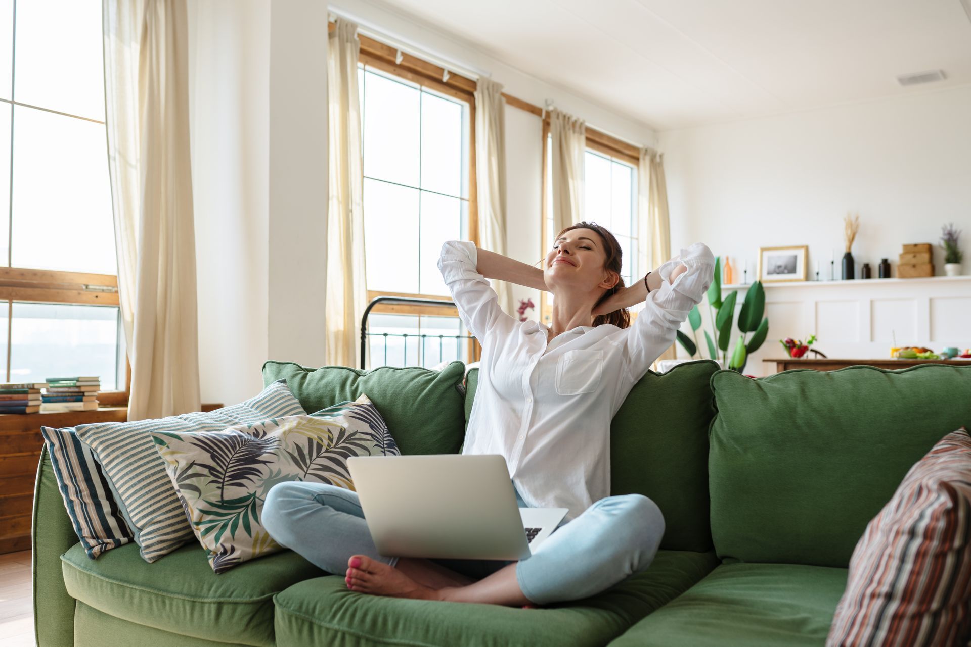 A woman is sitting on a couch with a laptop and stretching her arms.