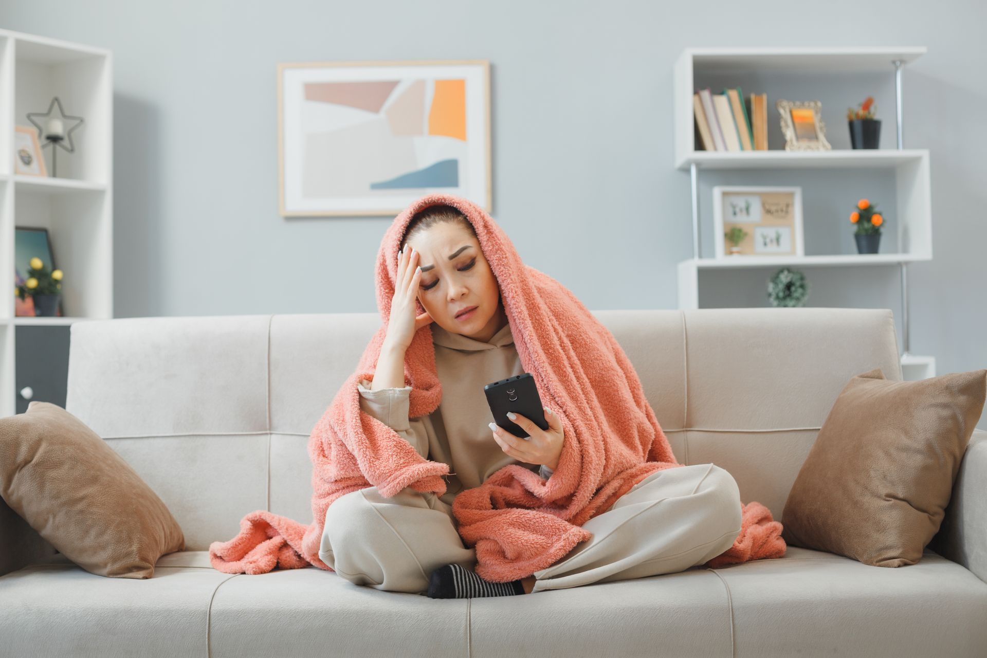 A woman wrapped in a blanket is sitting on a couch looking at her phone.