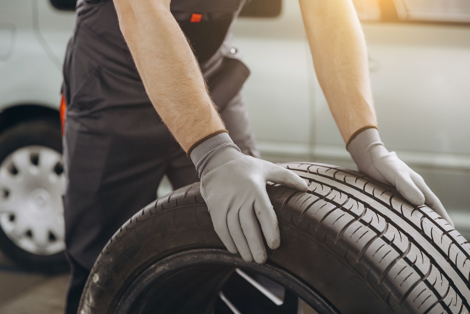Mechanic in grey gloves holding a tire, near a vehicle.