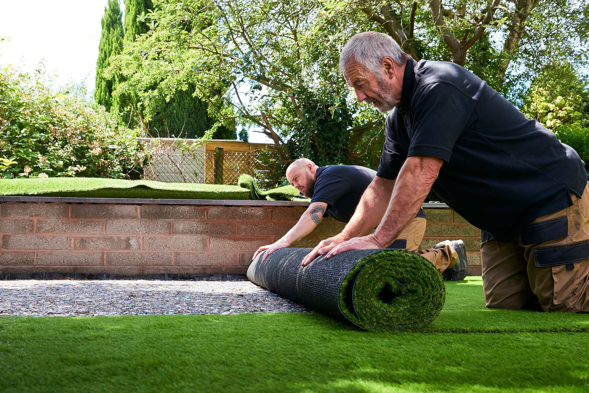 Two men are rolling a roll of artificial grass in a garden.