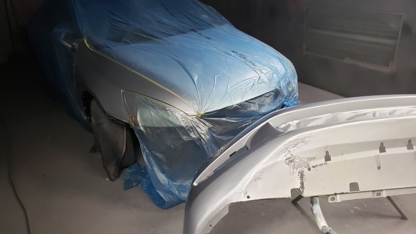 Blue car covered with a tarp in a garage, with a white car bumper in the foreground