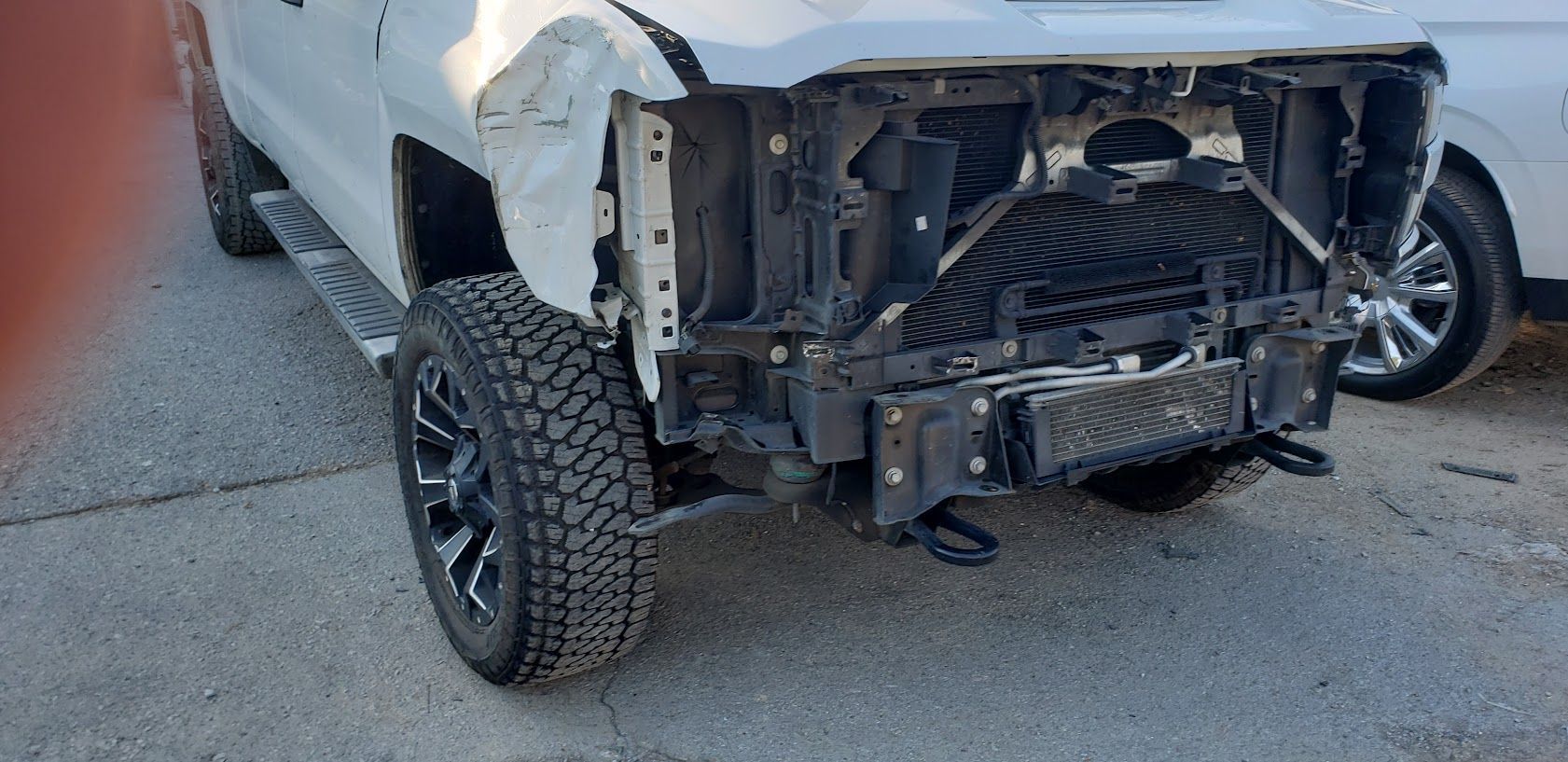 Silver truck with severe front-end damage, missing bumper, parked on a road.