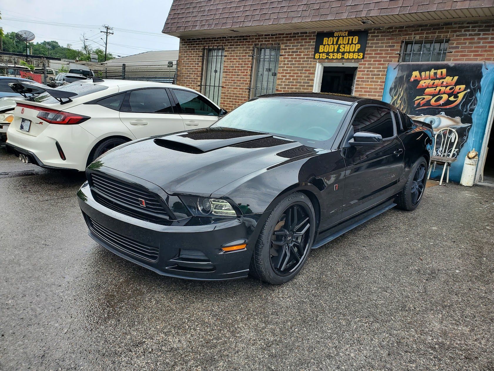 Black sports car parked outside a brick shop on a wet lot, with a white car beside it.