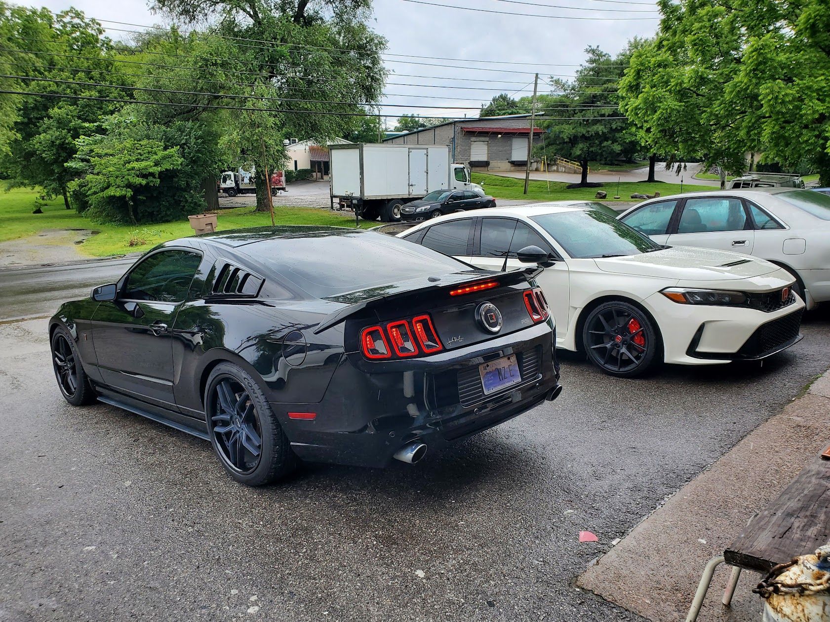 Three parked sports cars on a wet driveway, including a black Mustang and a white Toyota with red accents.