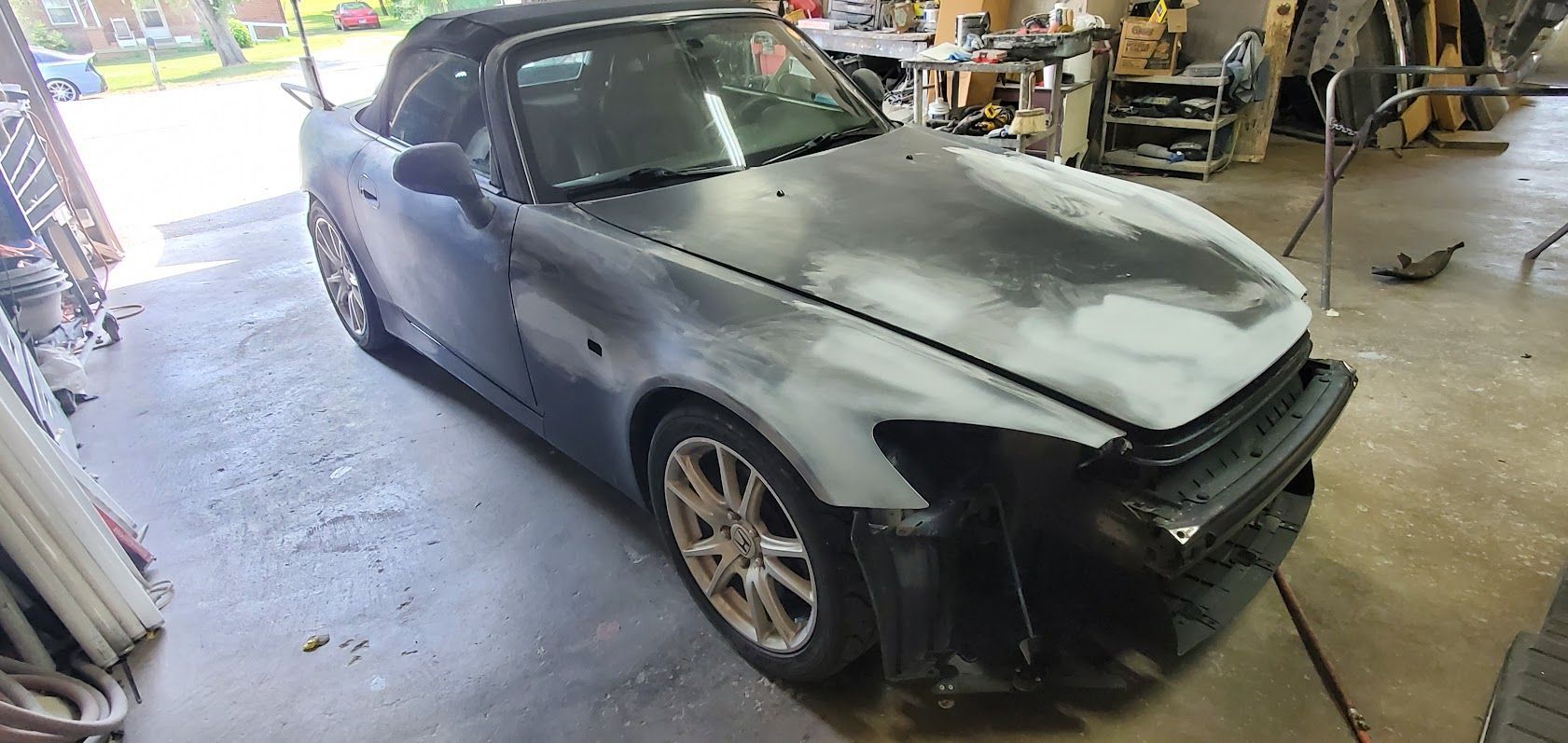 Matte black car in an auto body shop, front bumper missing and hood primed for repair