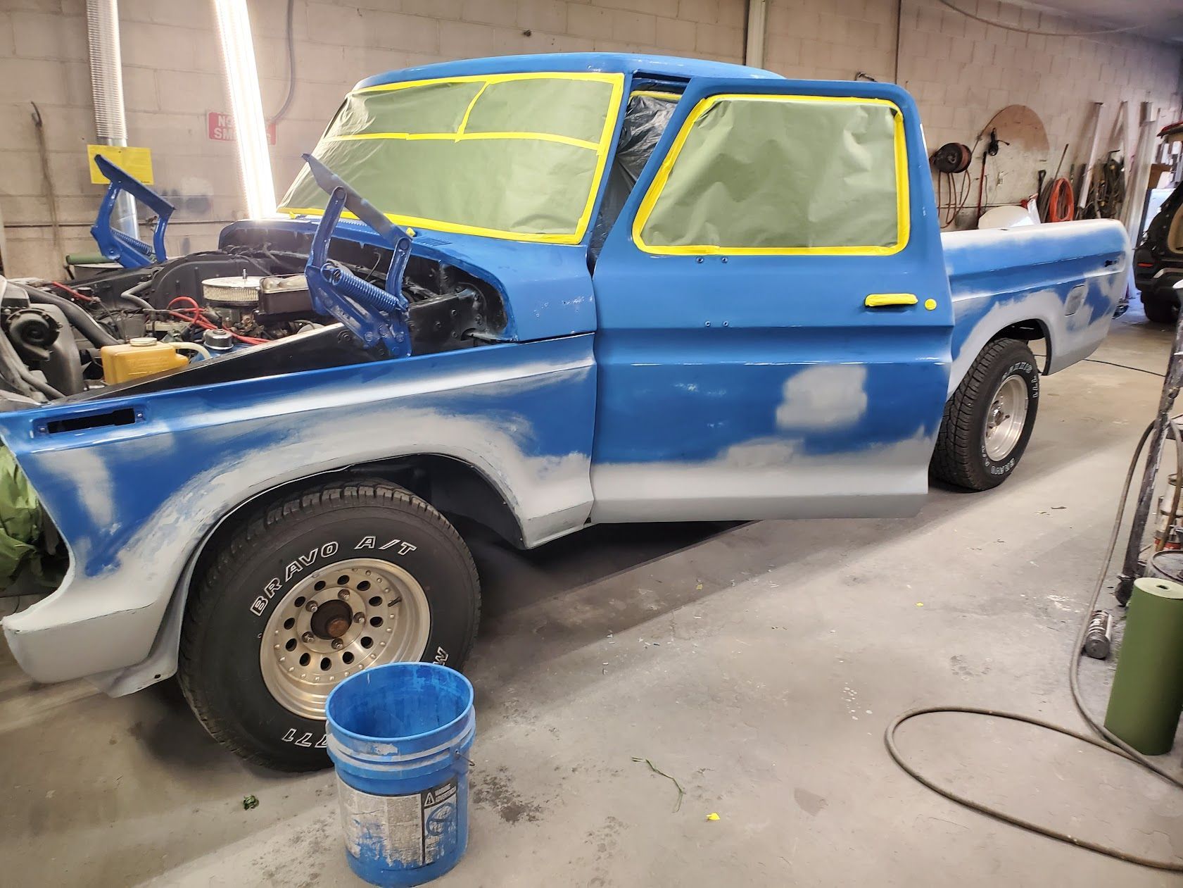 Blue pickup truck in a garage, partially primed with yellow-taped windows and hood removed.
