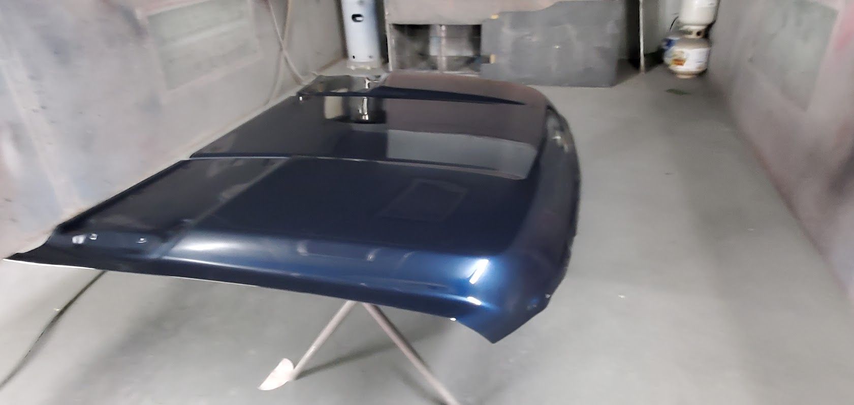 Blue car hood propped open in a spray booth or workshop, with a white stand beneath it