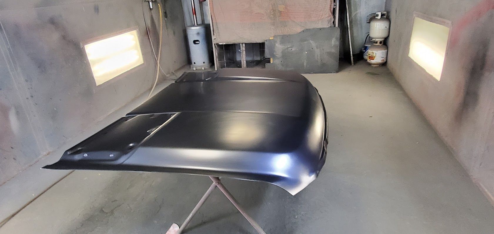 Black vehicle body panel inside a spray booth with white walls and small windows