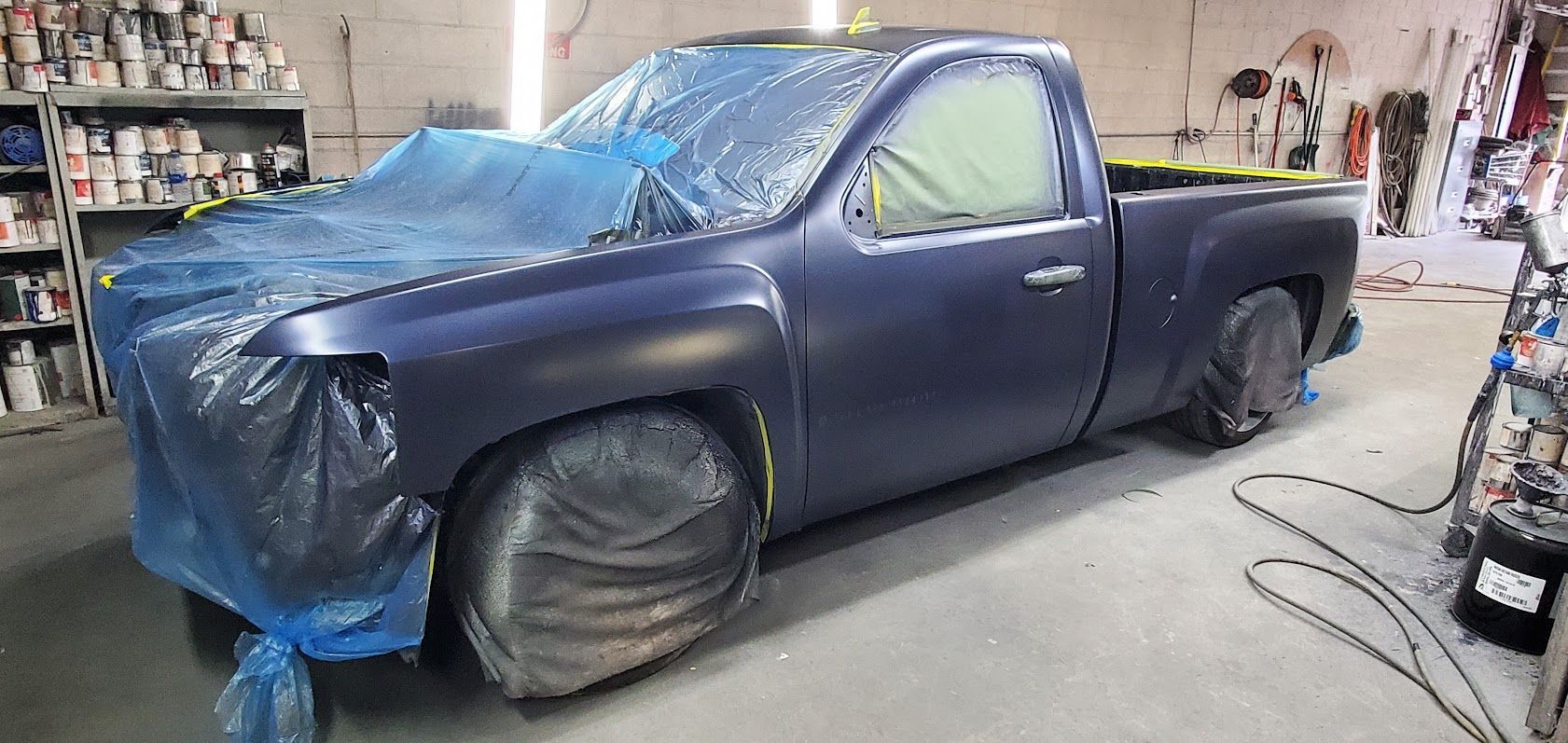 Blue pickup truck wrapped in plastic in a workshop, with front bodywork removed and wheels covered