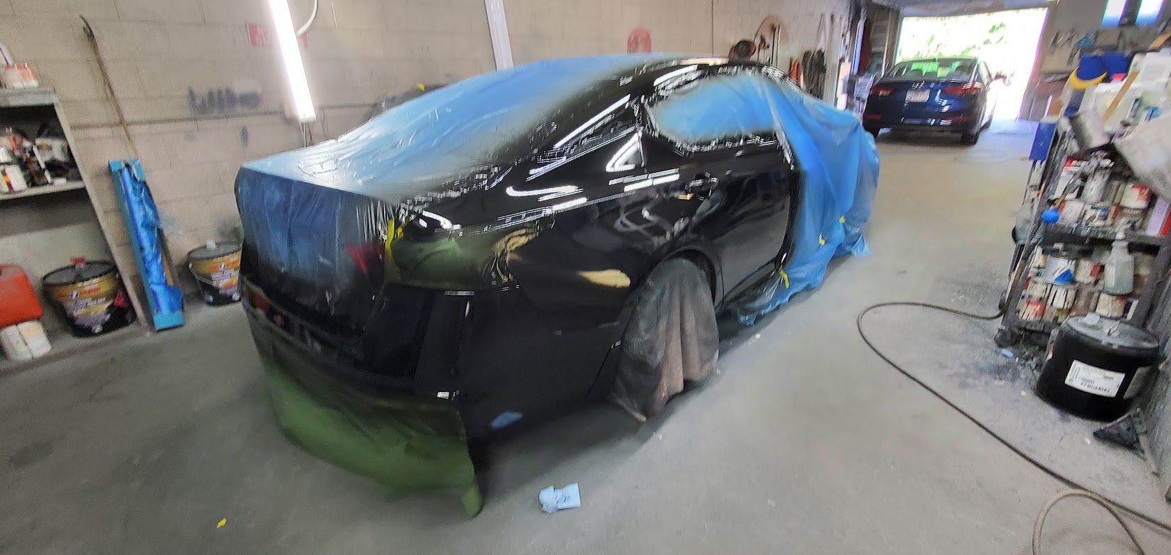 Black car in a workshop, partially covered with blue plastic and green masking for paint repair