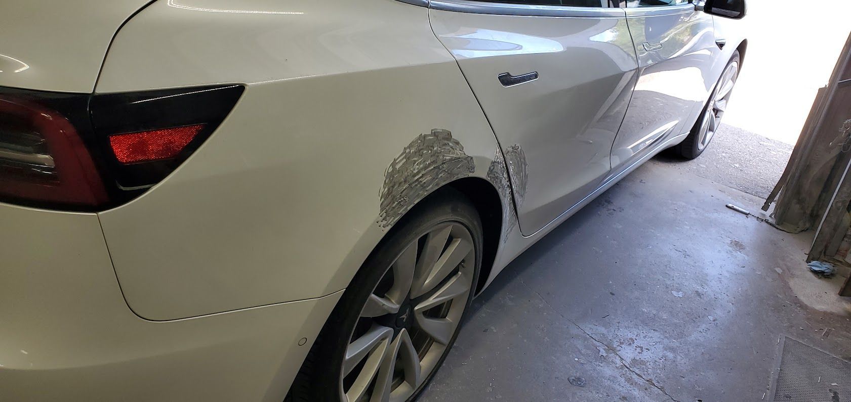 White sedan parked in a garage, rear side visible with dented right rear quarter panel.