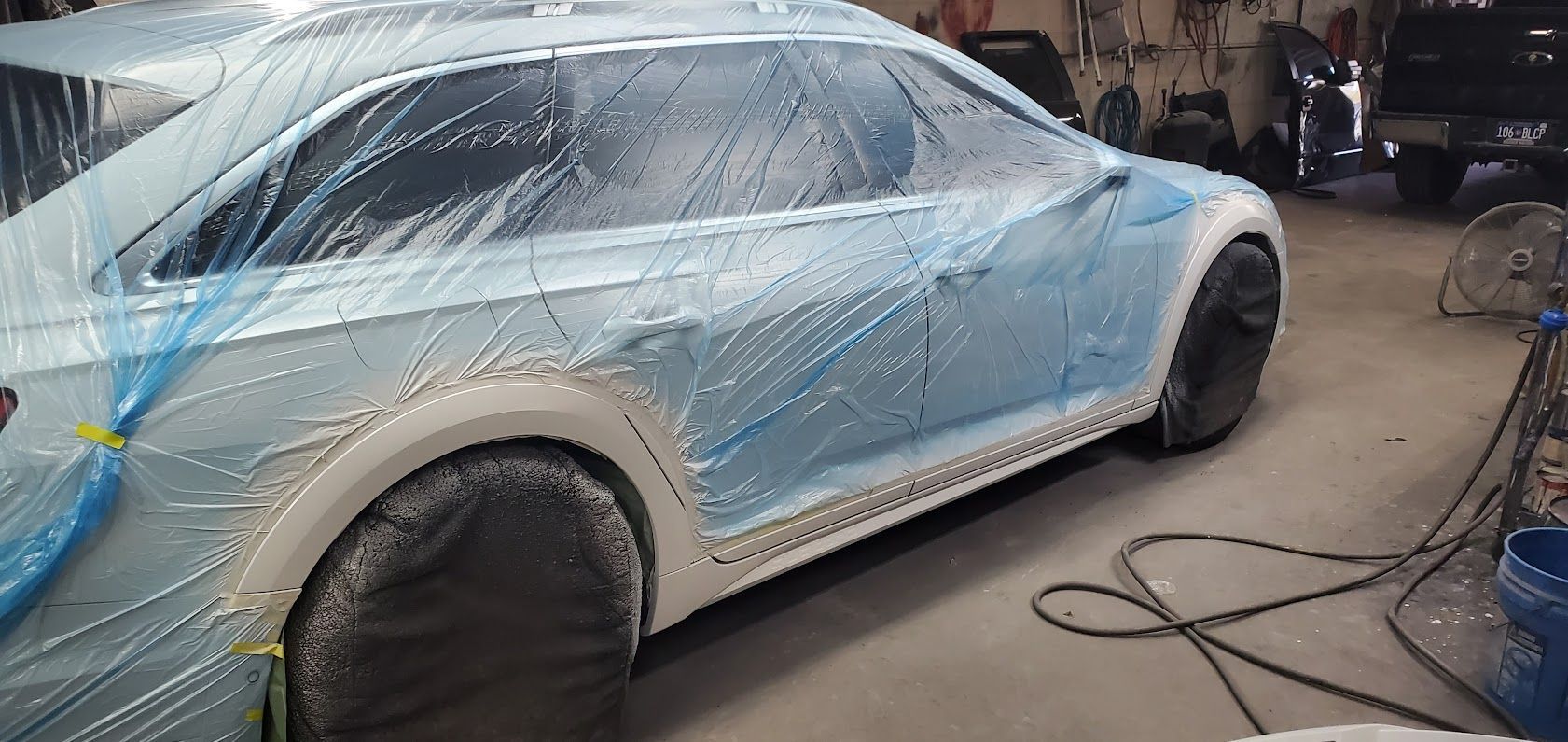 Blue car wrapped in plastic and masking tape in a workshop, with tires and body partially prepped for paint.