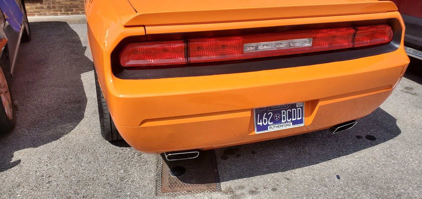 Rear view of an orange sports car with a black taillight bar and blue license plate, parked on asphalt.