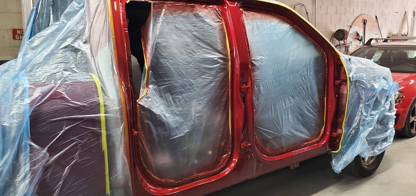 Red car body wrapped in plastic, with red trim and exposed doors in a workshop.