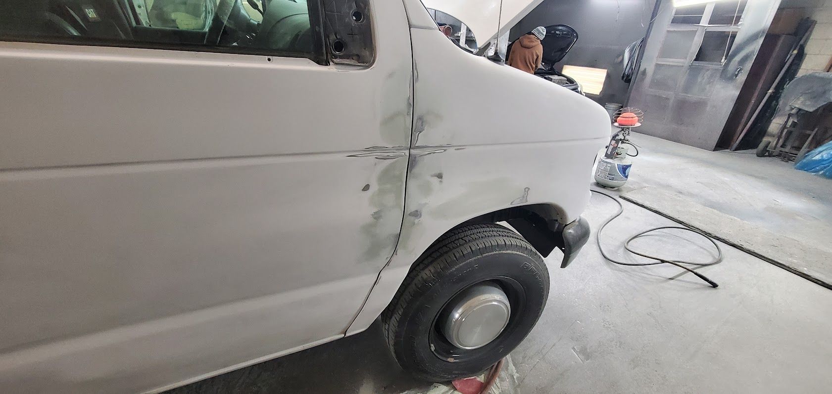 White van with a damaged front side panel and wheel, parked in a garage or repair area