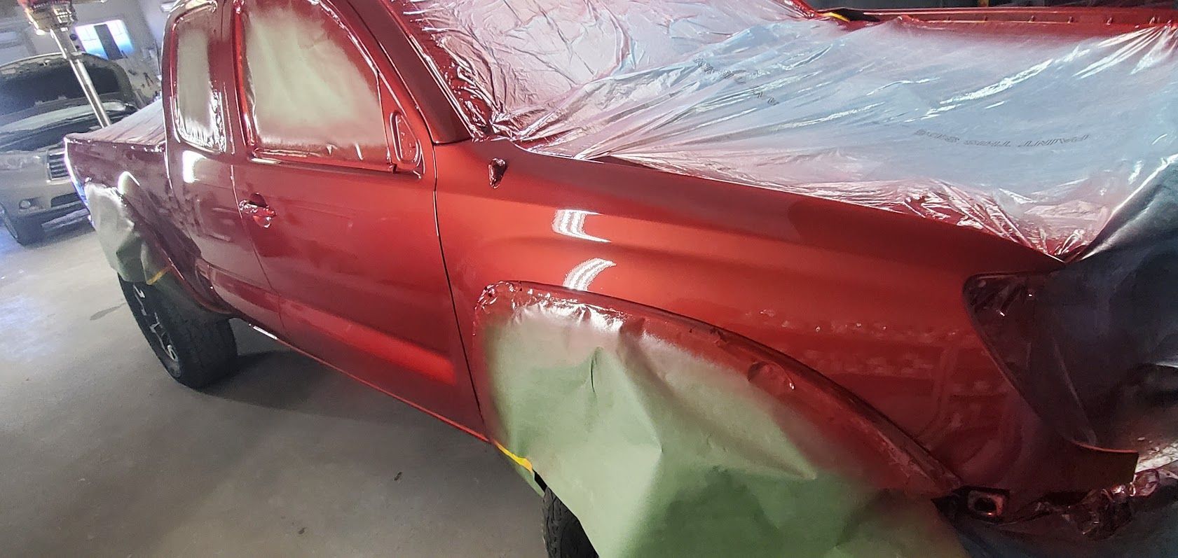 Red pickup truck with crumpled side and dented front, covered with protective plastic in a workshop