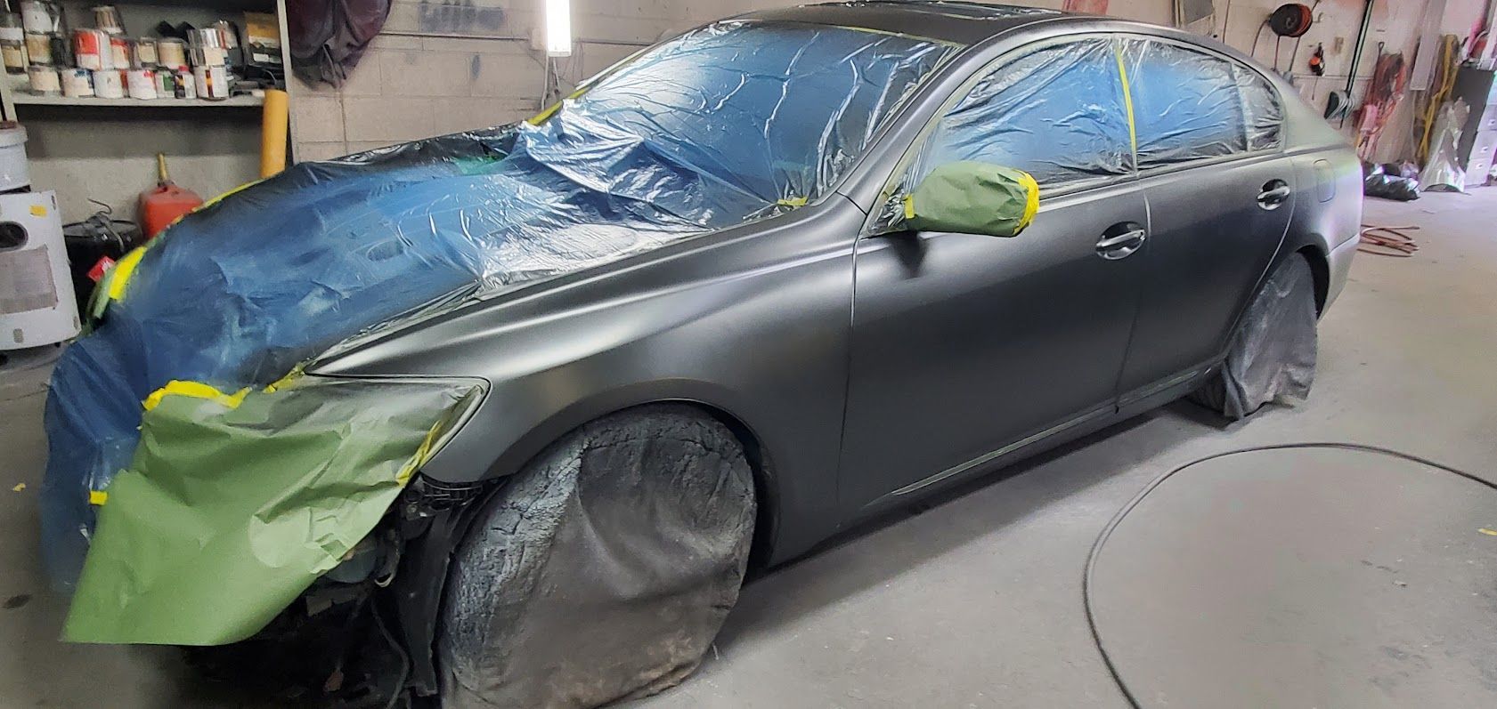 Blue sedan in a body shop, partially masked with green tape and plastic, awaiting paint work
