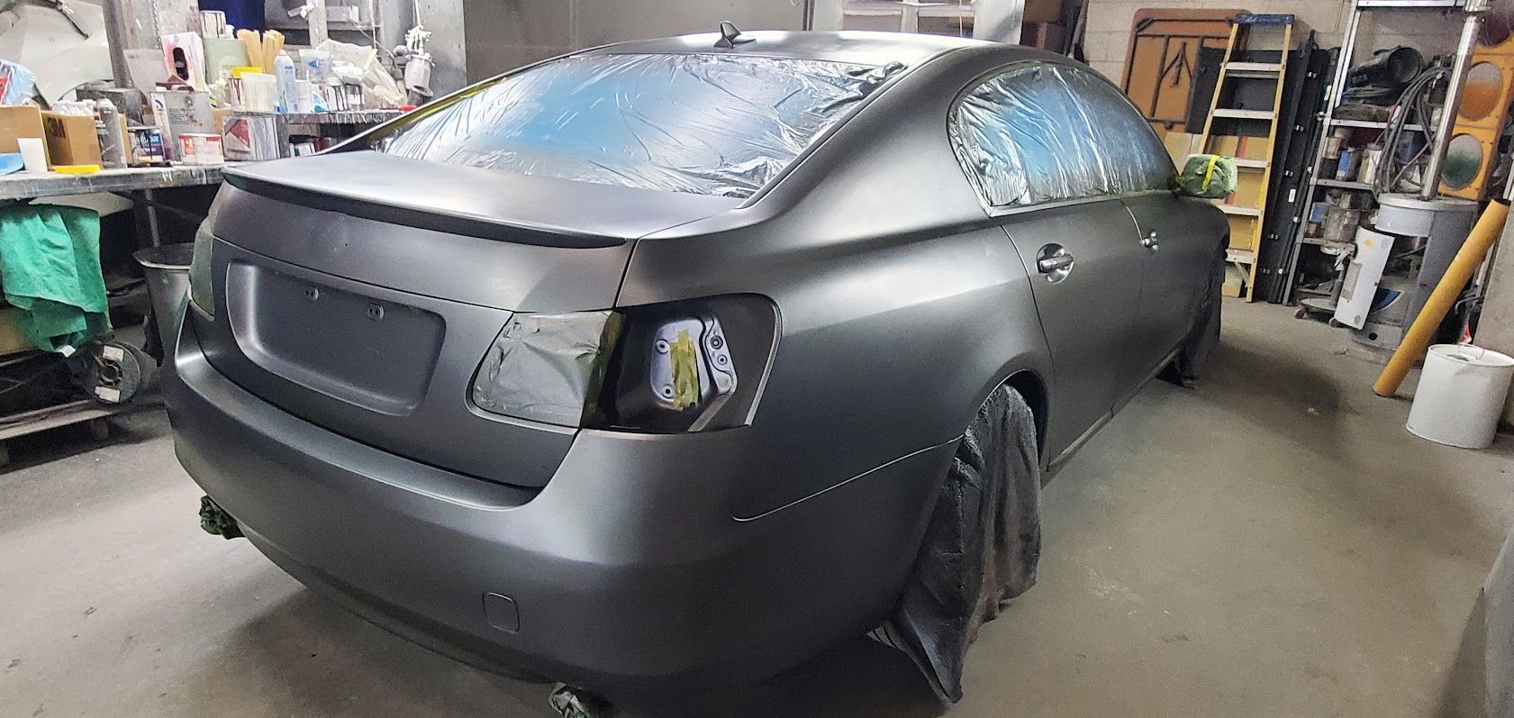 Matte black sedan in a garage, front-left view, with workshop shelves and tools in the background