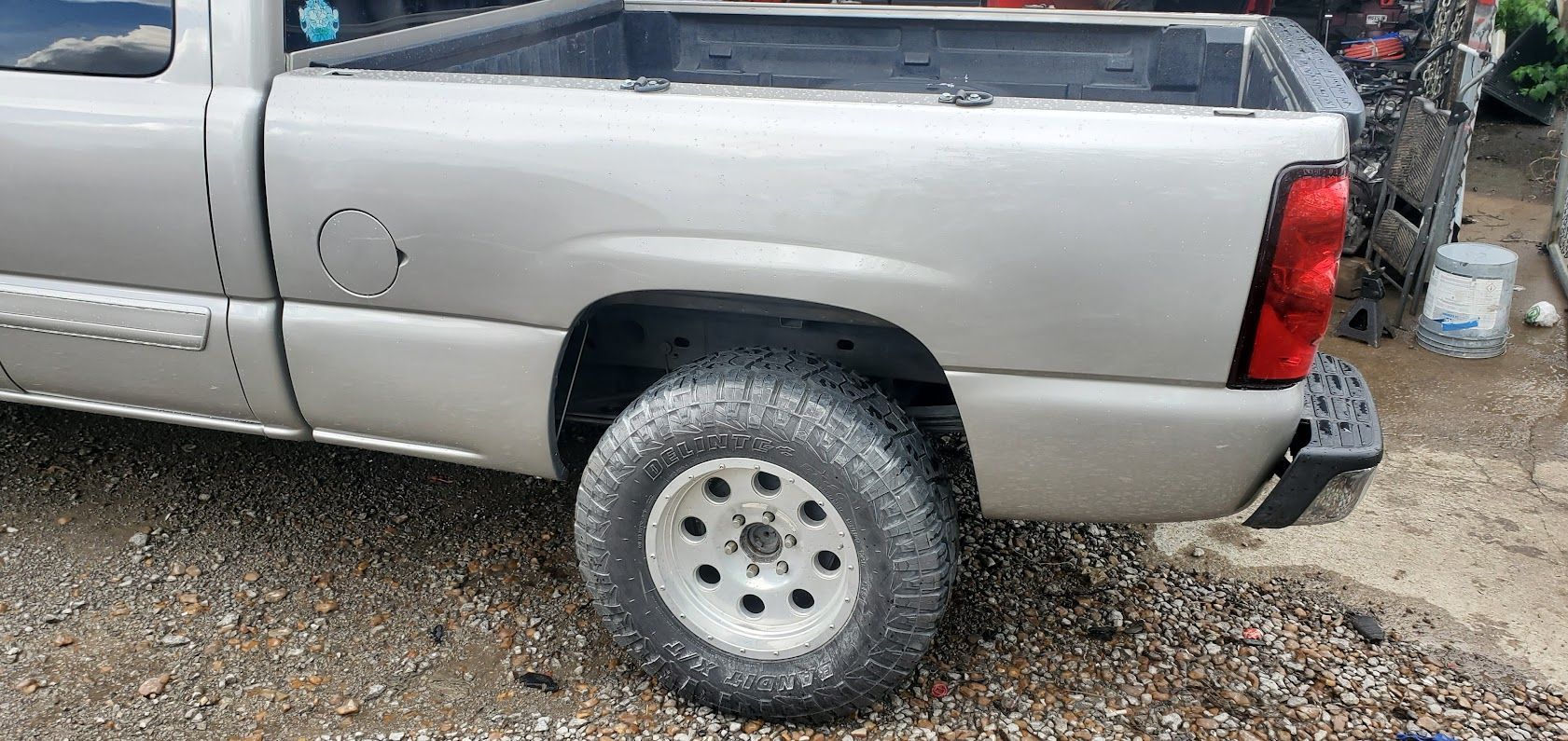 Silver pickup truck parked on gravel, rear side view with visible wheel and taillight