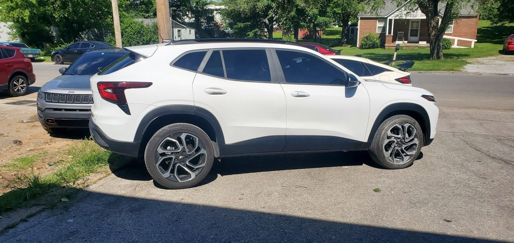 White SUV parked on a sunny residential street, side view, with a red car and trees in the background.