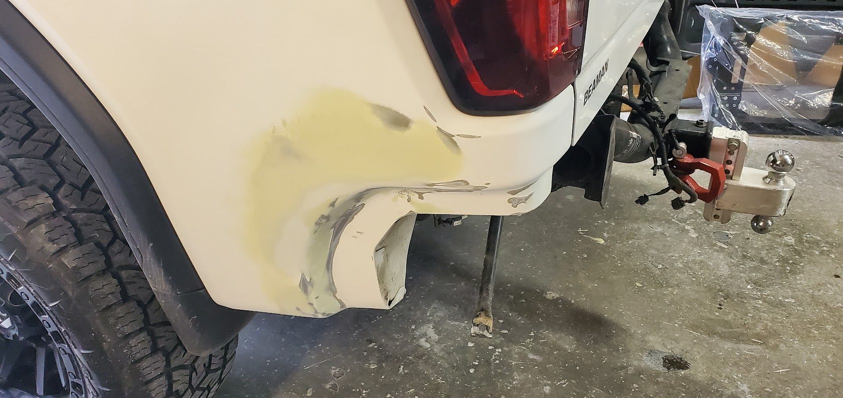 White vehicle rear bumper with damaged yellow paint and a trailer hitch in a workshop.