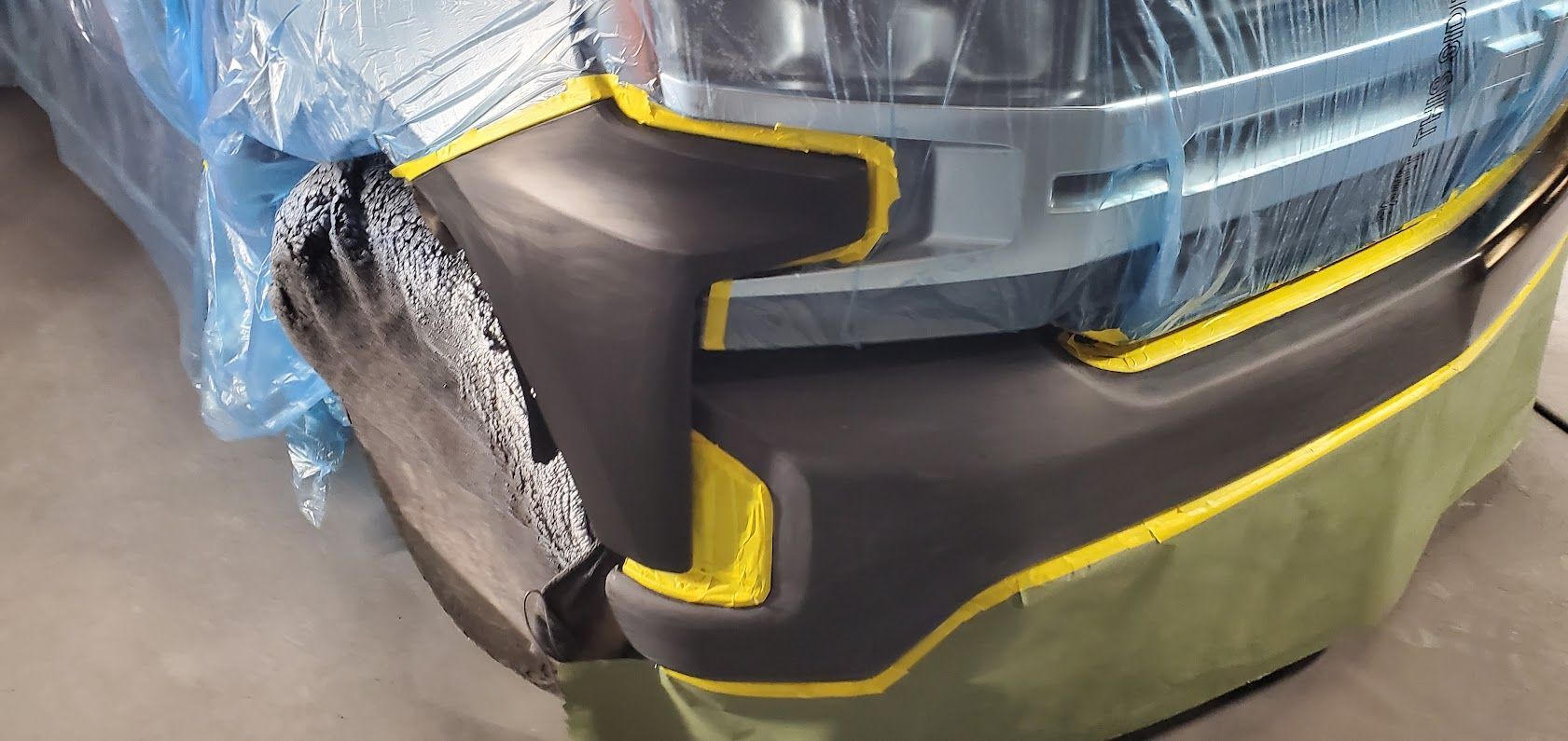 Close-up of a black and yellow car bumper wrapped in blue protective plastic, in a workshop.