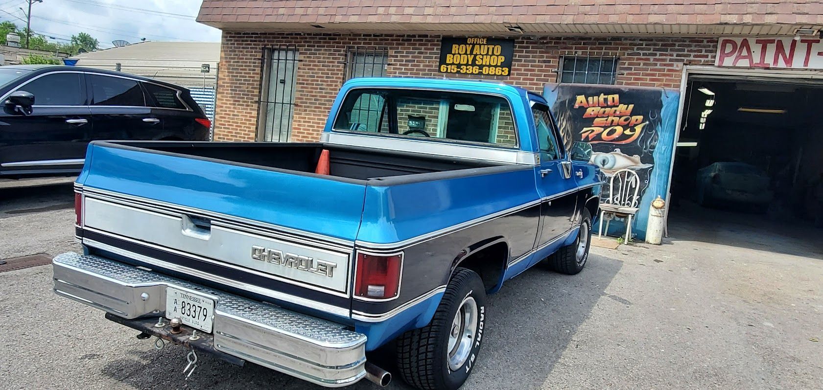 Blue classic pickup truck parked outside a rustic auto shop with a paint sign and open garage doors