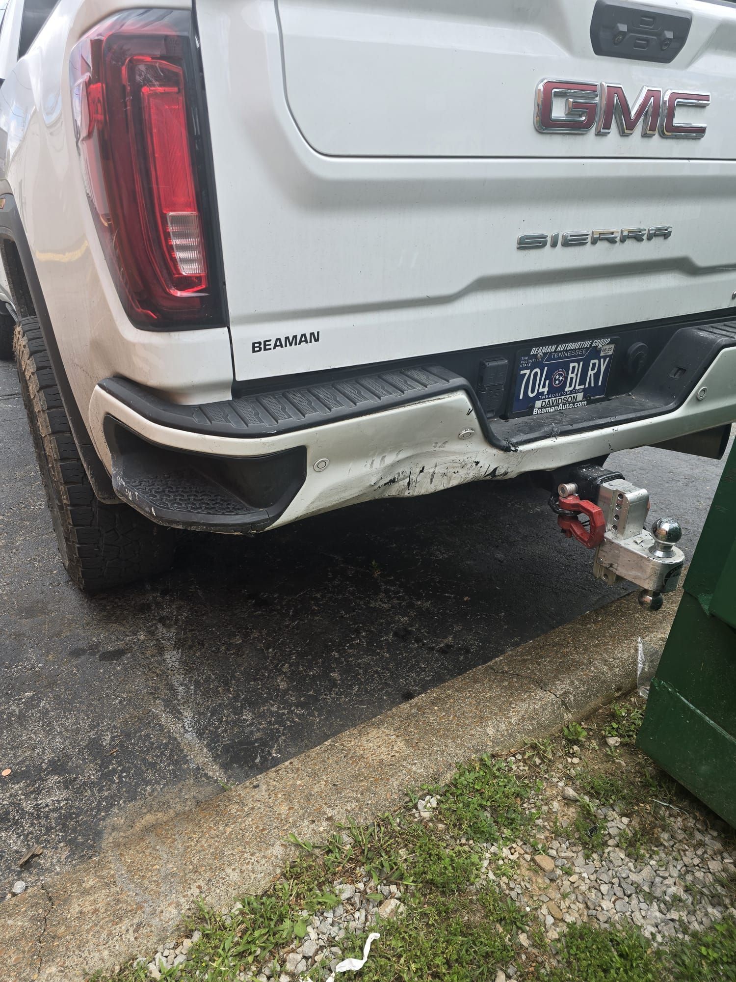 Rear of a white GMC SUV with bumper damage, parked by a curb with a tow hitch attached.