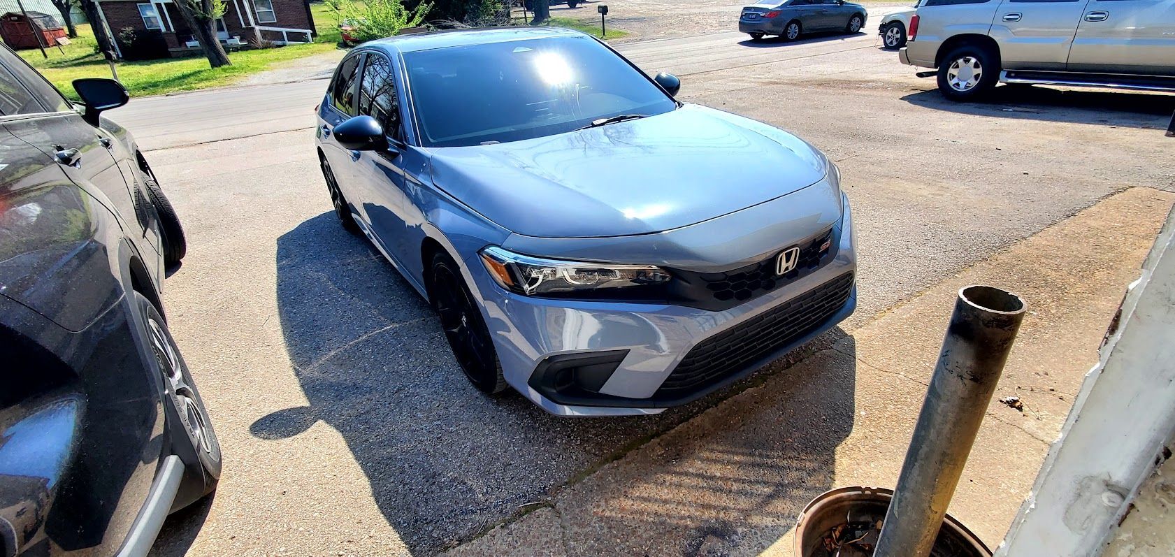 Silver sedan parked in a sunlit lot, front view with blue car and posts at the edges.