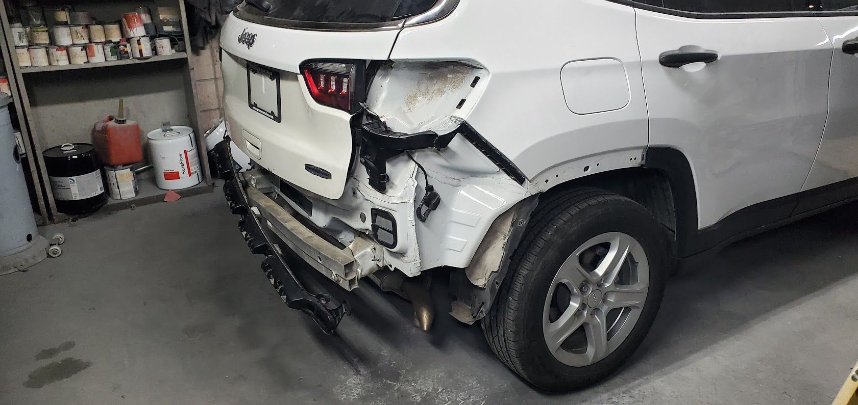 Damaged white SUV with rear-end and side impact in a garage, bumper hanging off.