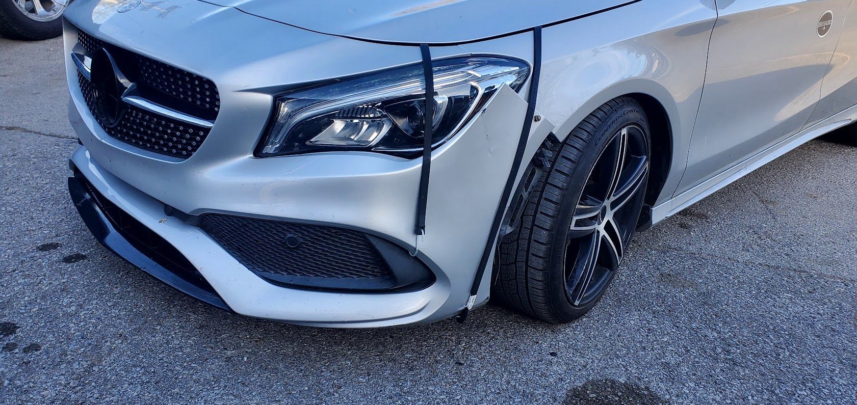 White Mercedes sedan with a damaged front bumper and bent wheel, parked on gravel.
